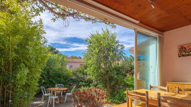 Un patio exterior con una mesa y sillas de madera, rodeado de abundante vegetación y una vista de una casa al fondo.