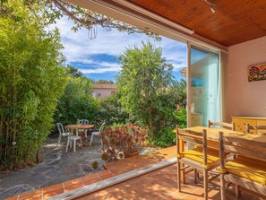An outdoor patio with a wooden table and chairs, surrounded by lush greenery and a view of a house in the background.