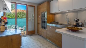Cocina moderna con armarios de madera, horno empotrado, microondas y un lavabo con grifo. Hay una puerta de vidrio que da a un patio con vistas a la vegetación.
