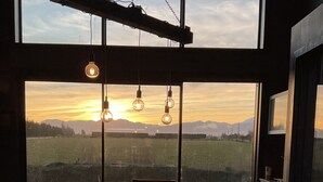 Un comedor con una mesa grande y sillas, un lámpara colgante, y vistas a un campo y montañas a través de ventanas amplias.