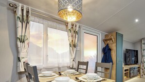 A dining area with a wooden table set for four, a chandelier, and a view of the outdoors through large windows. - Deluxe Plus Sierra 4 (Hunstanton)