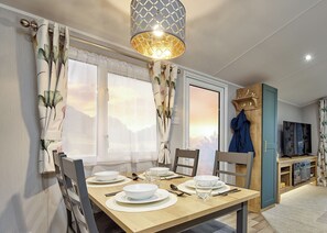 A dining area with a wooden table set for four, a chandelier, and a view of the outdoors through large windows. - Deluxe Plus Sierra 4 (Hunstanton)