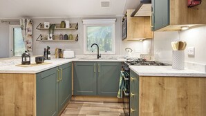 A modern kitchen with a large island, built-in appliances, and a window with a view of greenery. - Deluxe Plus Sierra 4 (Hunstanton)