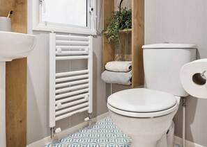 A bathroom with a toilet, a white radiator, a wooden shelf with towels, and a window. - Deluxe Plus Sierra 4 (Hunstanton)