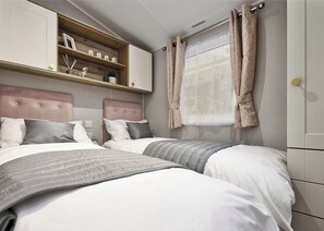 A bedroom with two beds, a wooden shelf with decorative items, a window with curtains, and a headboard. - Deluxe Plus Sierra 4 (Hunstanton)