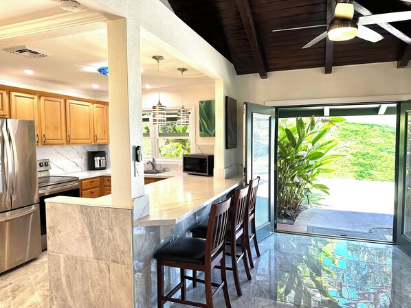 A modern kitchen with a large island, stainless steel appliances, wooden cabinets, and a view of greenery outside. - Newly Remodelled 4-bedroom cottage in Amazing Kaanapili with ACs (Lahaina)