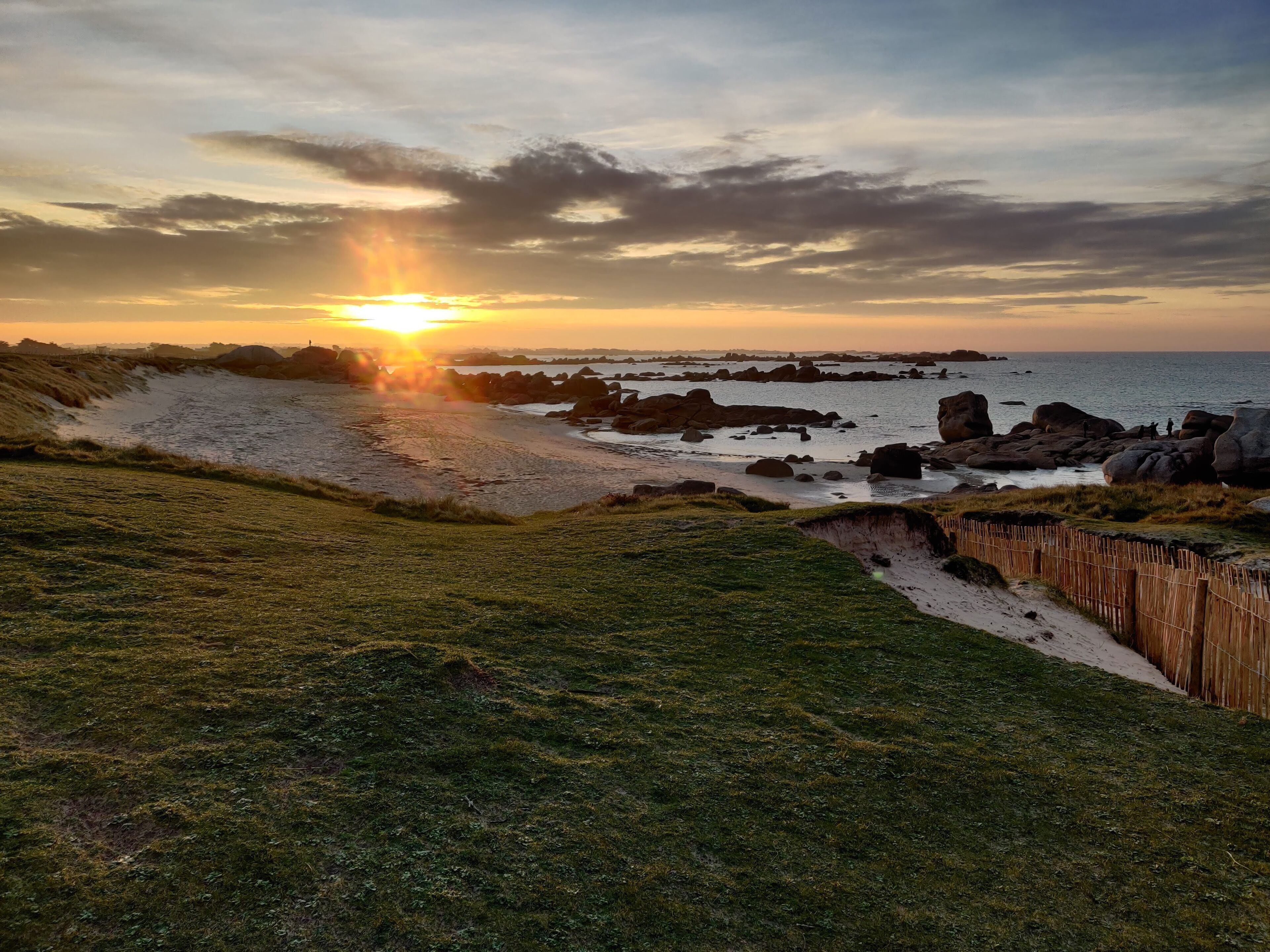 A coastal landscape with a sandy beach, grassy areas, and a wooden fence.