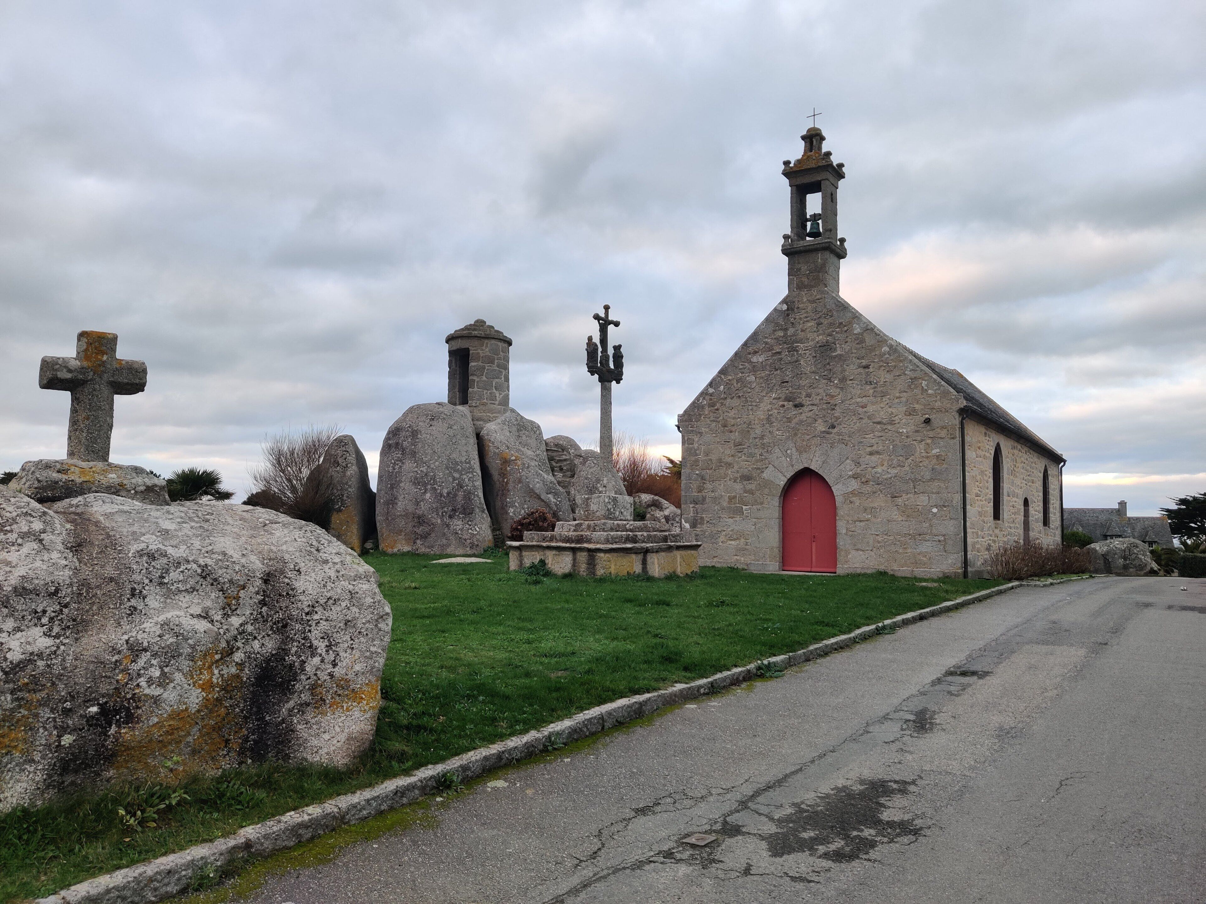 Uma igreja de pedra com uma porta vermelha, grandes pedras e uma cruz em primeiro plano.