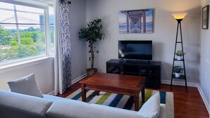 A living room with a sofa, a coffee table, a TV, a ceiling fan, a window with curtains, a plant, and a painting on the wall.