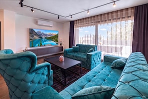 Un salón moderno con un sofá grande, dos sillones y una mesa de centro. Un cuadro de un paisaje montañoso adorna la pared, y hay un ventanal con cortinas.