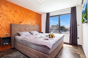 A modern bedroom with a large bed, wooden wall paneling, a window with curtains, and a flat-screen TV.