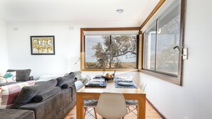 Een eetgedeelte met een houten tafel en stoelen, een grijze bank en een groot raam met uitzicht op bomen.