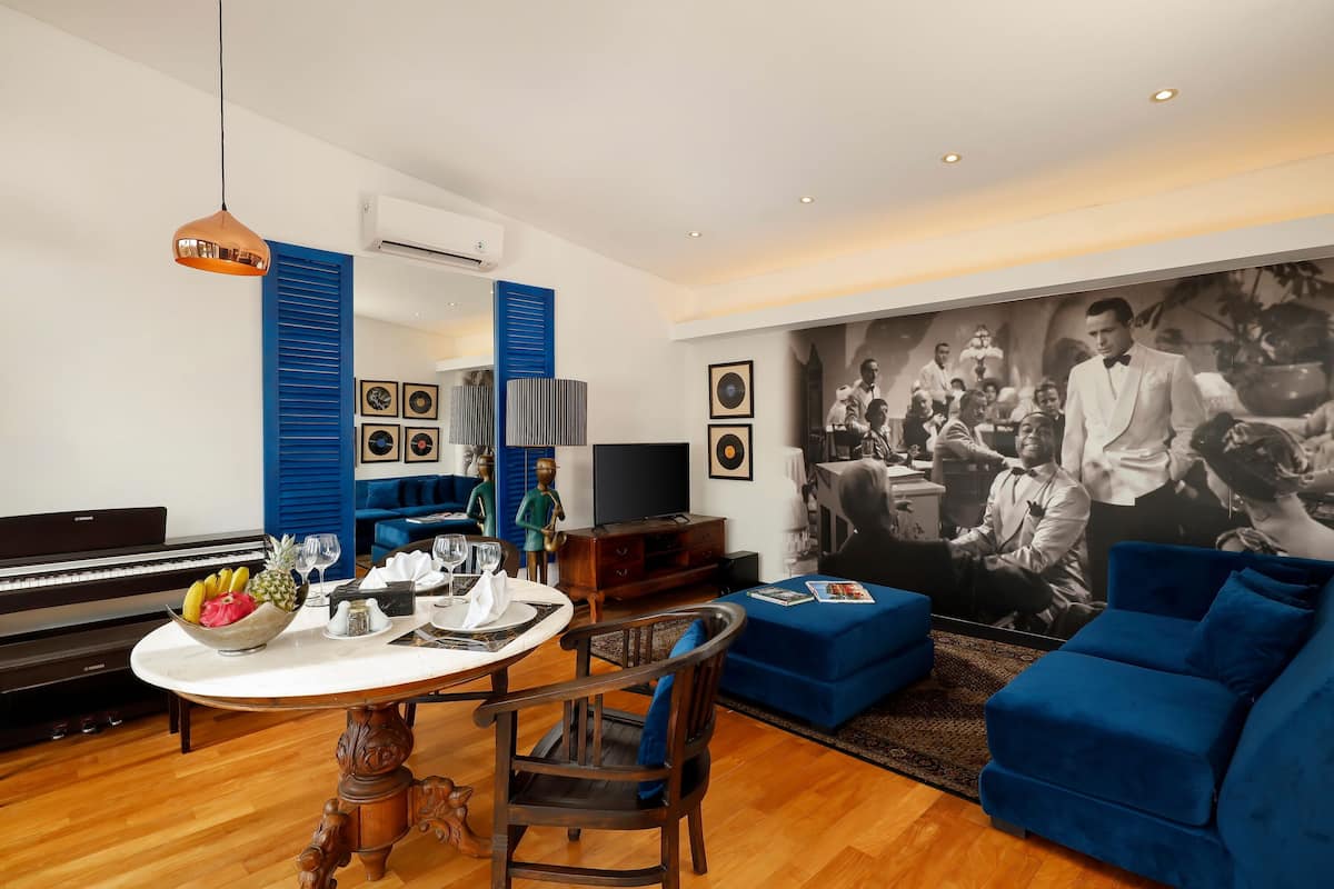 Un salon avec un piano, une table à manger ornée de fruits et de verres, un canapé bleu et une grande photographie en noir et blanc accrochée au mur.