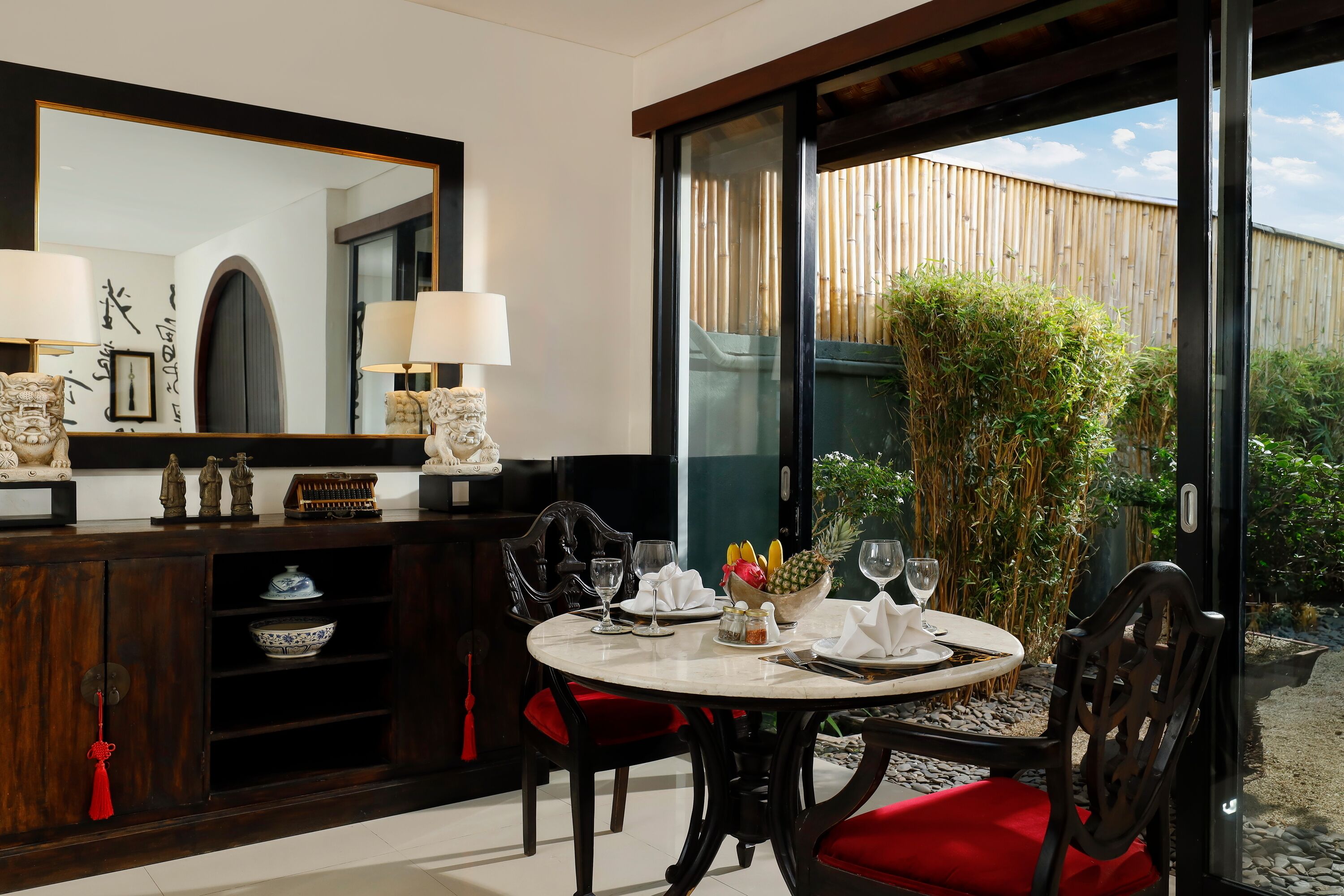 A dining area with a round table, red cushioned chairs, and a large window offering a view of bamboo and greenery.
