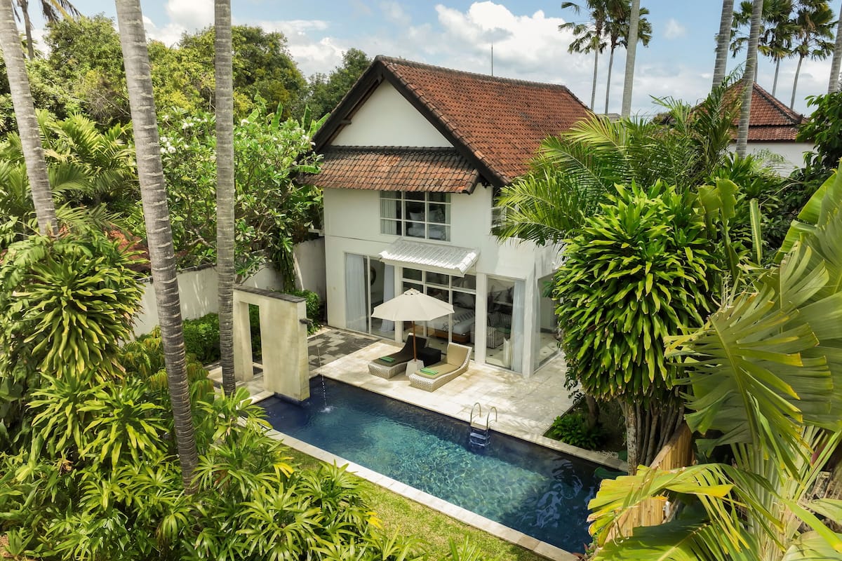 Une maison à deux étages avec une piscine, entourée d’une végétation luxuriante et de palmiers.