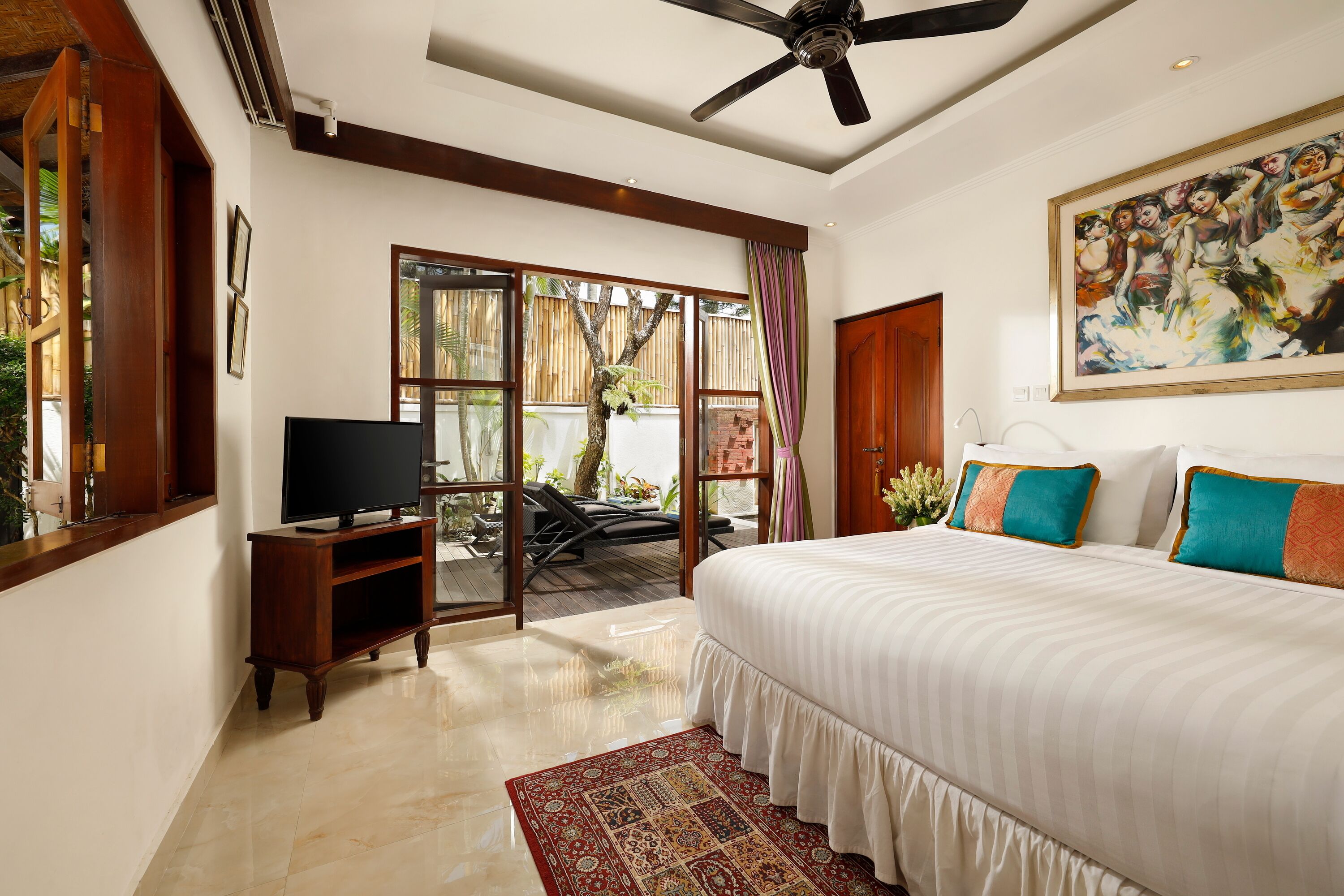 A bedroom with a large bed, a television on a wooden stand, a ceiling fan, and a sliding glass door leading to an outdoor area.