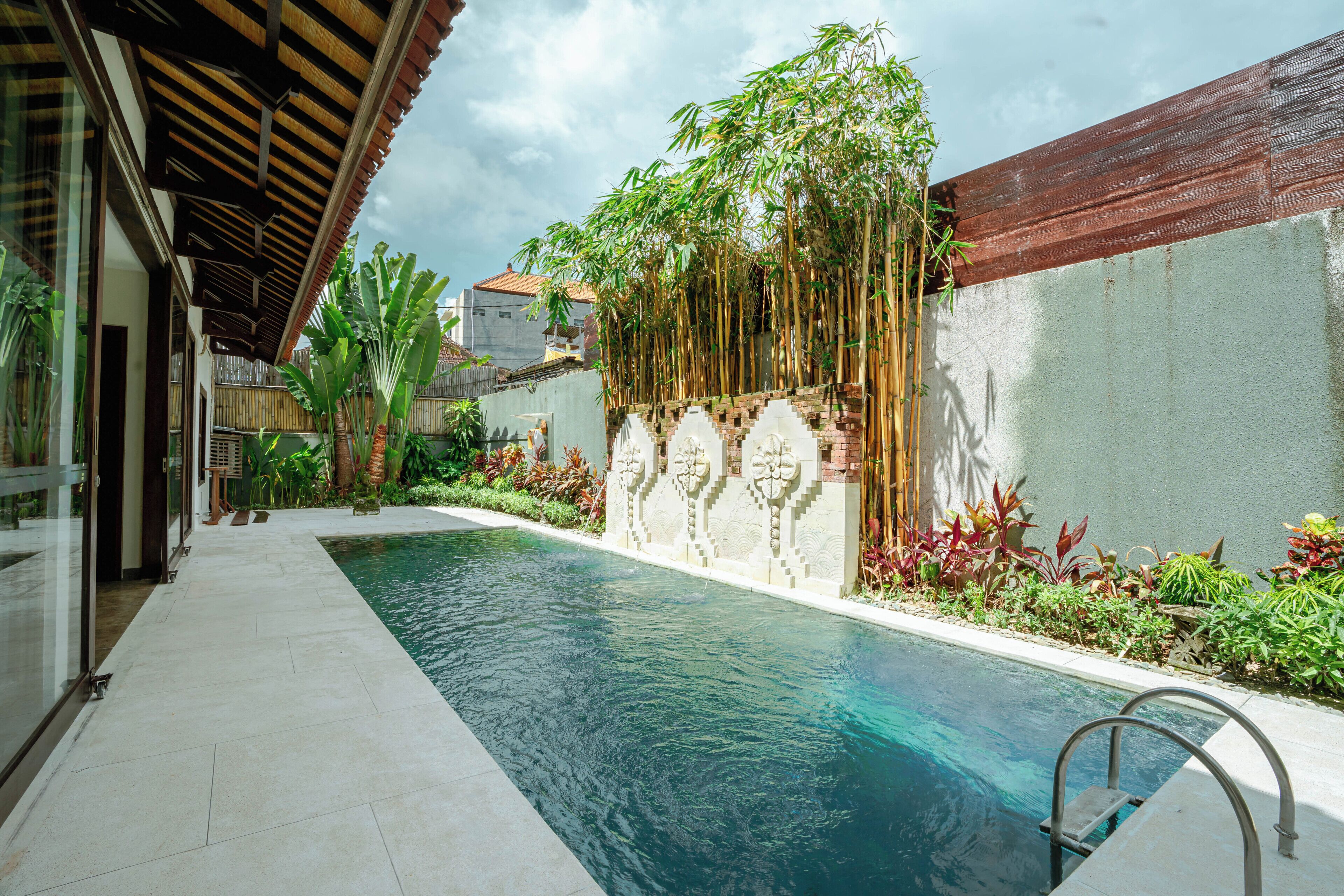Una piscina con bordo piastrellato, circondata da vegetazione lussureggiante e un muretto decorativo con piante di bambù verticali.