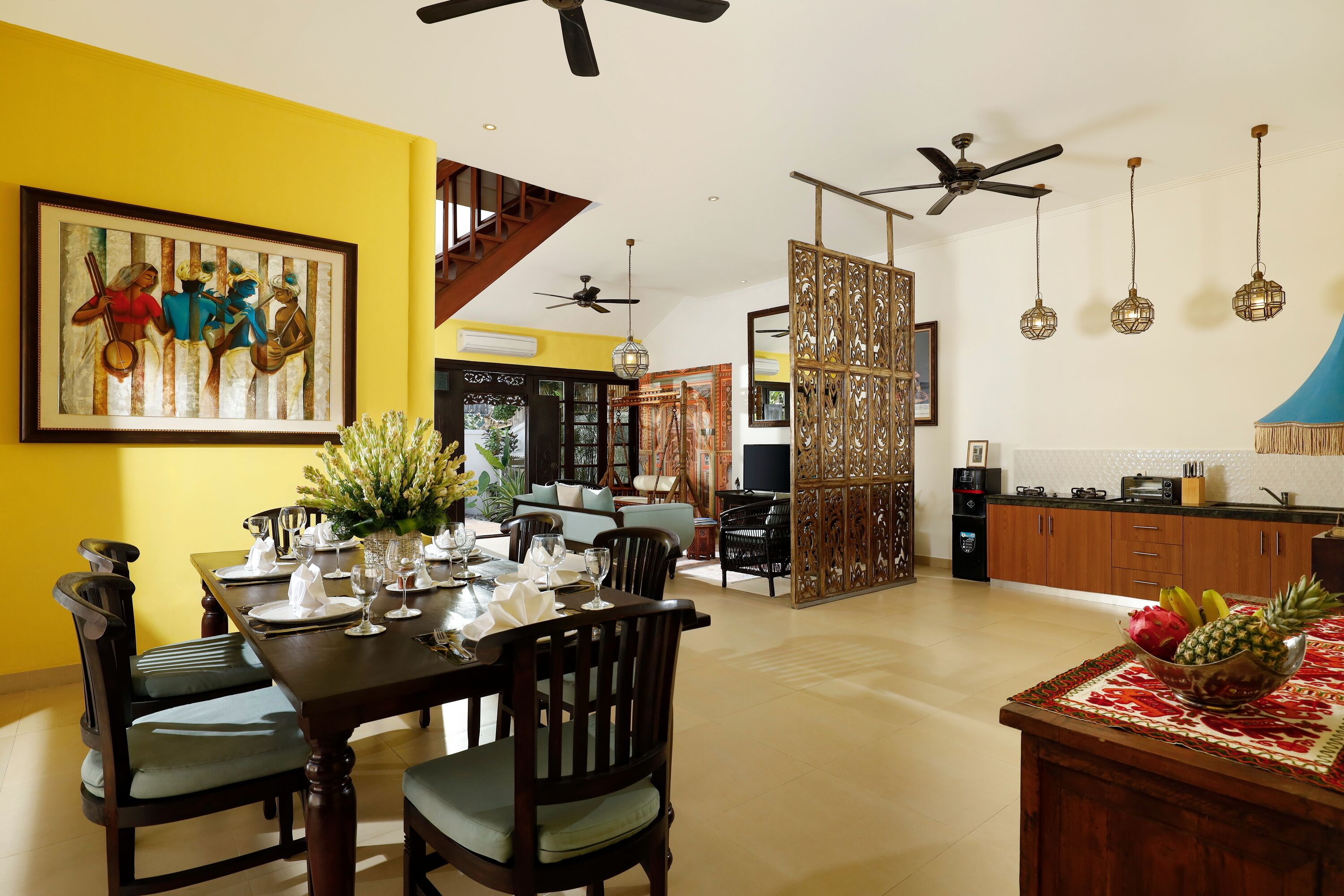Uma sala de jantar com uma mesa de madeira posta para quatro pessoas, uma cozinha ao fundo e uma escada que leva a um andar superior.