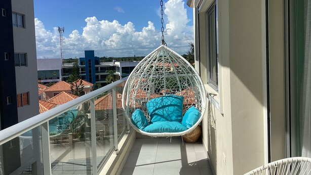 A balcony with a hanging chair, glass railing, and a view of buildings and a clear sky.