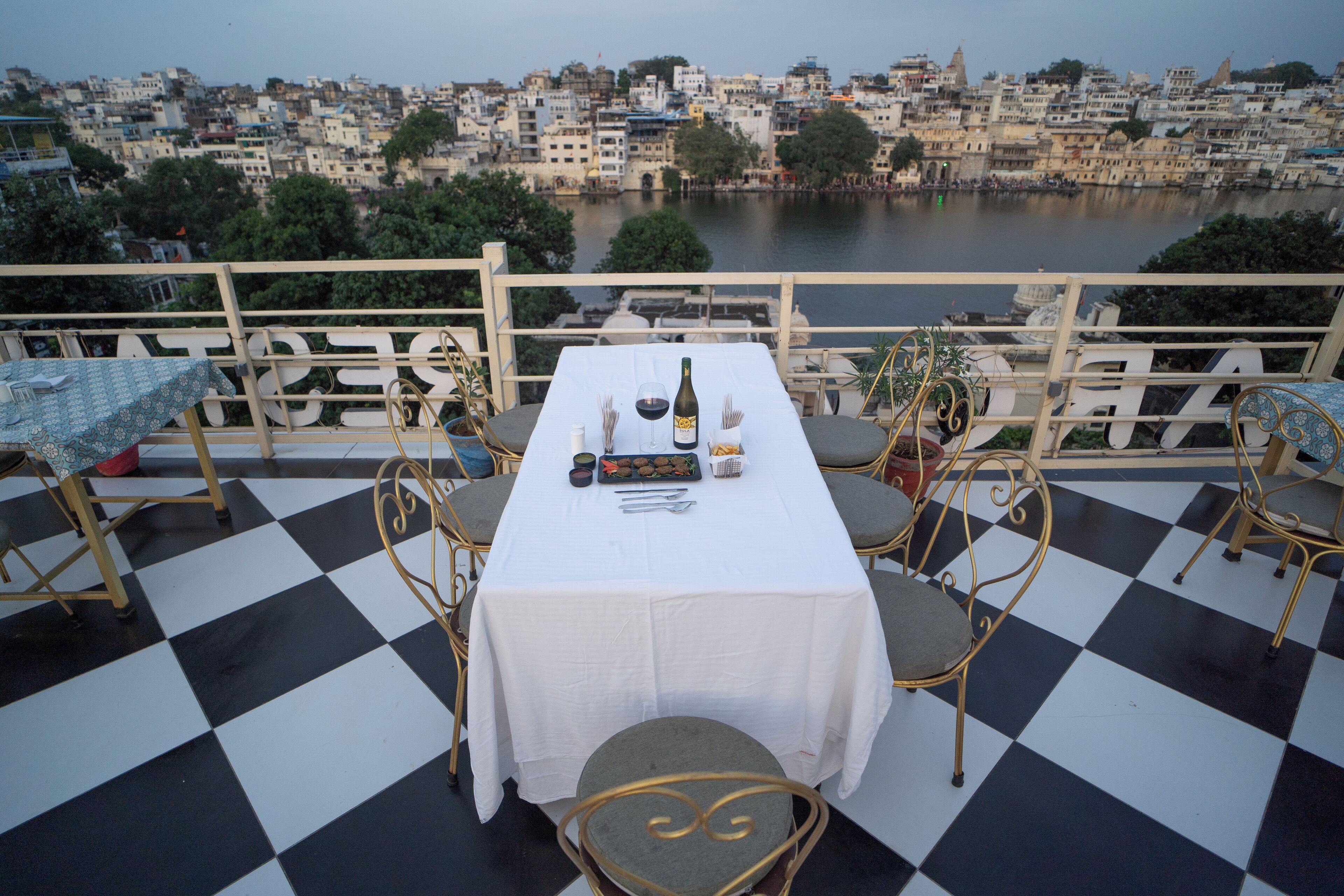 Une table dressée avec une bouteille, des verres et des amuse-gueules sur une terrasse dont le sol est à carreaux et qui est équipée de chaises en métal.