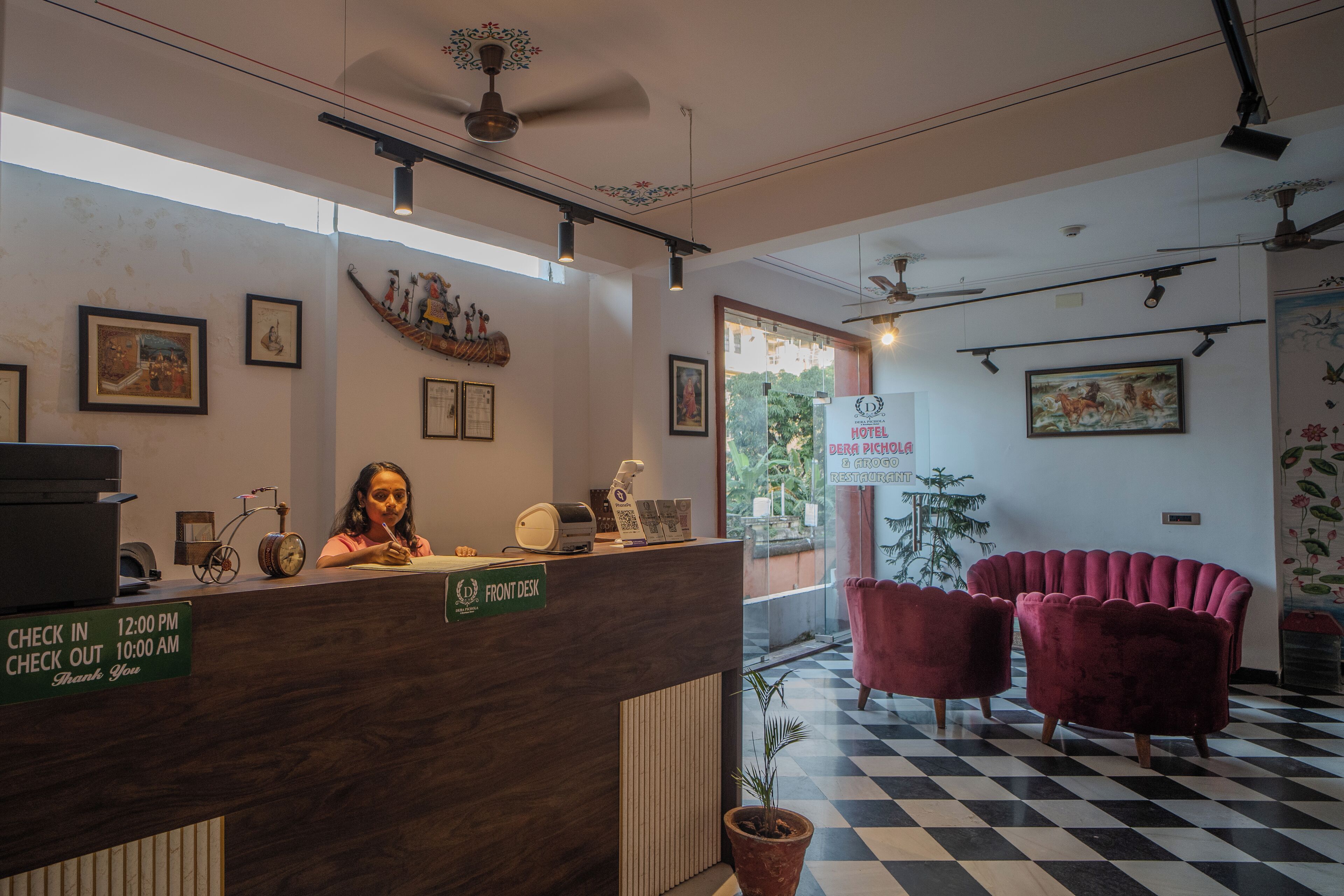 A hotel reception area with a check-in/check-out desk, framed artwork on the walls, and a check-in/check-out sign.