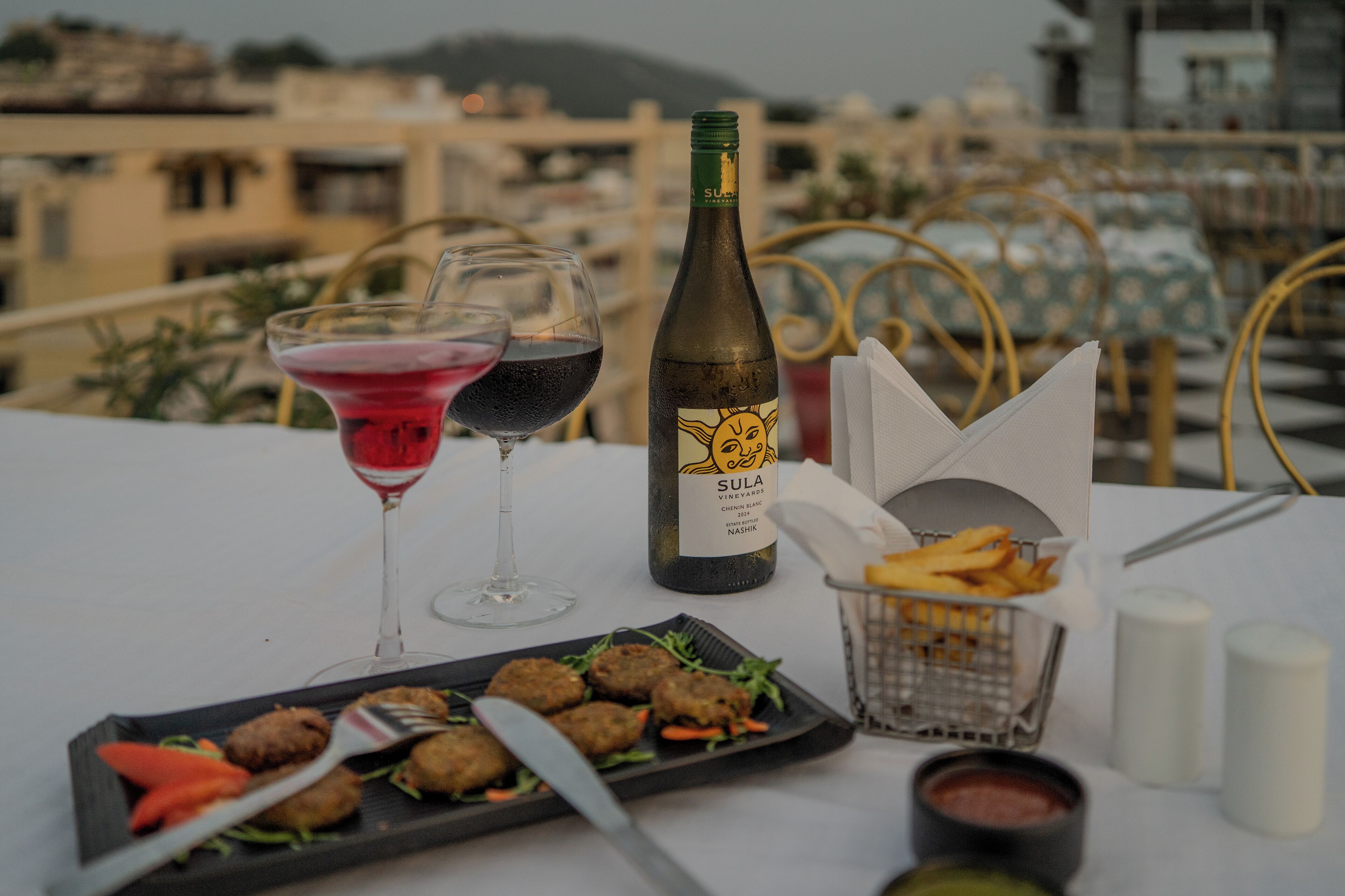 Una mesa con una botella de vino Sula Vineyards, dos copas de vino tinto, un plato de comida, una cesta de papas fritas y salsas.