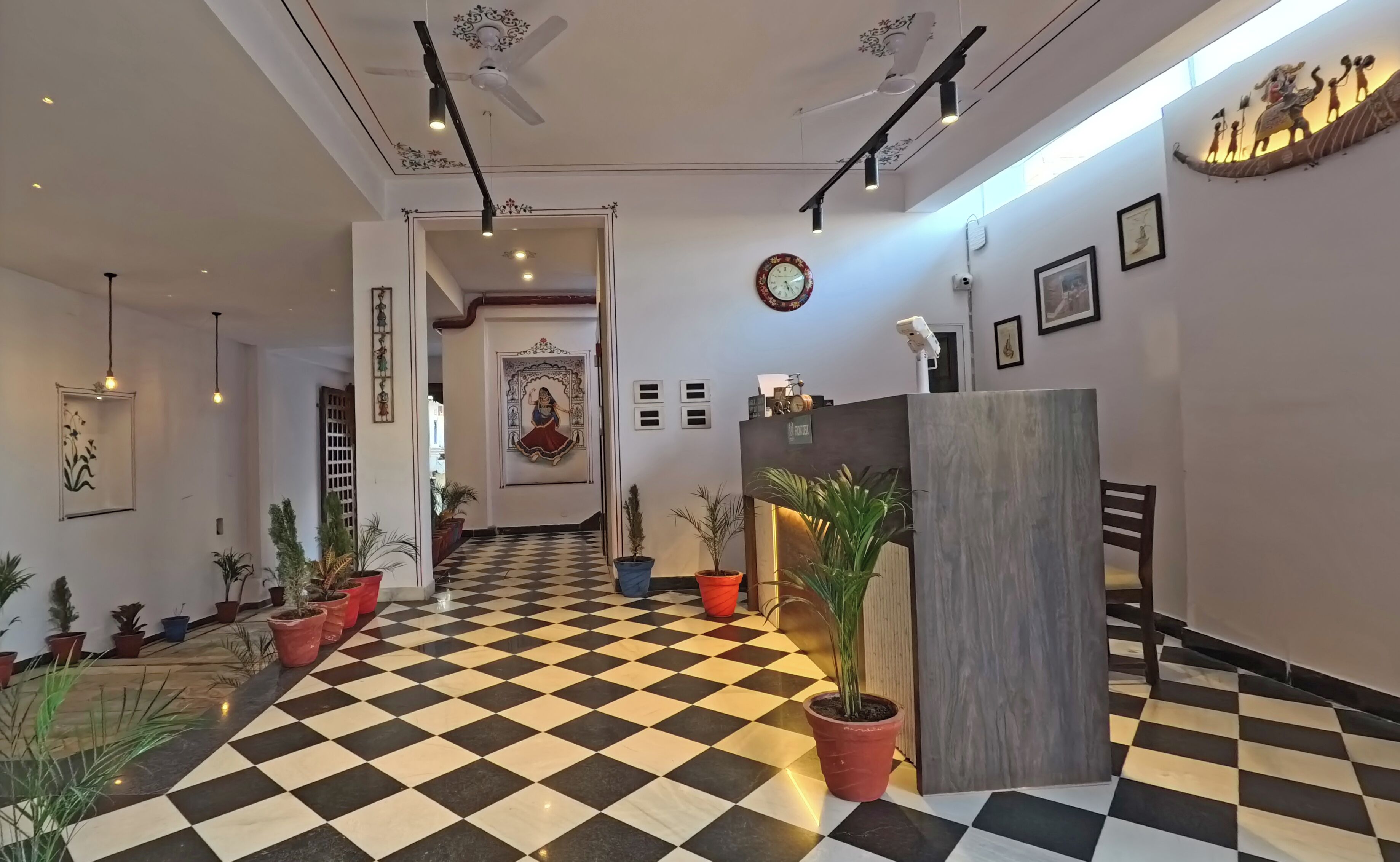 Un hall de hotel con piso a cuadros, ventiladores de techo, lámparas fijadas a la pared, plantas en macetas, una recepción y un elemento decorativo en la pared.