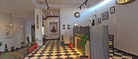 Un hall de hotel con piso a cuadros, ventiladores de techo, lámparas fijadas a la pared, plantas en macetas, una recepción y un elemento decorativo en la pared.
