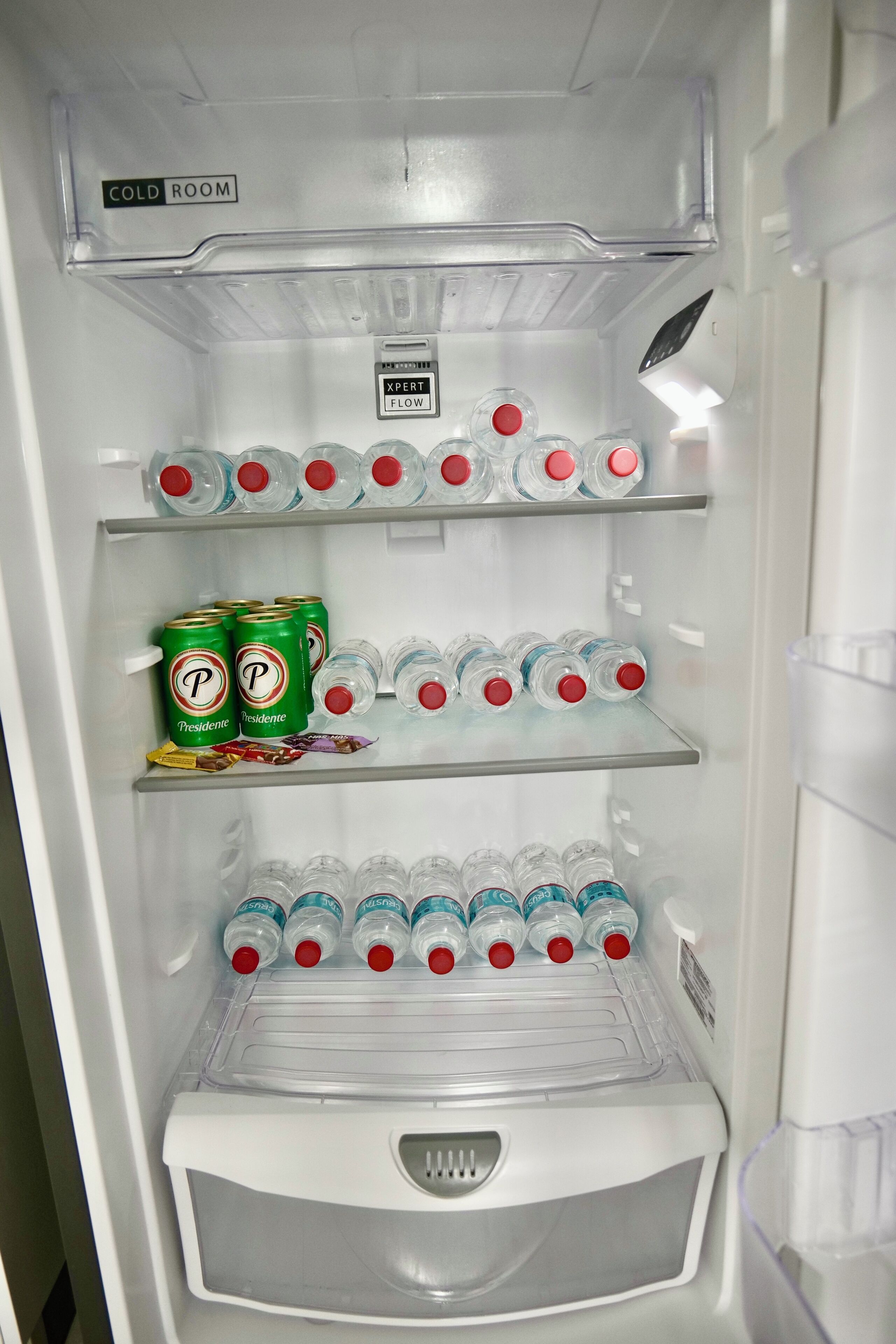 A refrigerator with a 'COLD ROOM' label, multiple shelves with bottled water, and cans of 'Presidente' beer.