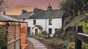A narrow stone pathway leading to a white house with a dark roof, surrounded by greenery and a wooden structure on the left. - The Coronation Family Cottage | Ridgestone (Lindale)