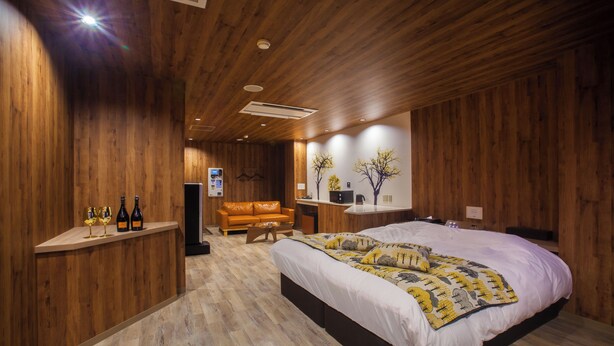 A wooden-paneled bedroom with a bed, a sofa, a desk, and a shelf with bottles and glasses. - HOTEL elLle - Adults Only (Toyama)