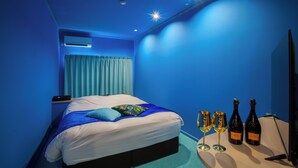 A bedroom with a bed, a nightstand, an air conditioning unit, a window with curtains, and two bottles of champagne on the floor. - HOTEL elLle - Adults Only (Toyama)