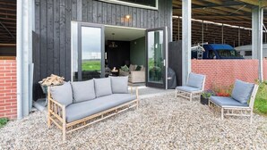 A modern outdoor seating area with a sofa and chairs, a gravel floor, and a building with a dark exterior and large glass doors. - Luxury Farmstay – Sleep in Style Among the Cows (Loenen aan de Vecht)
