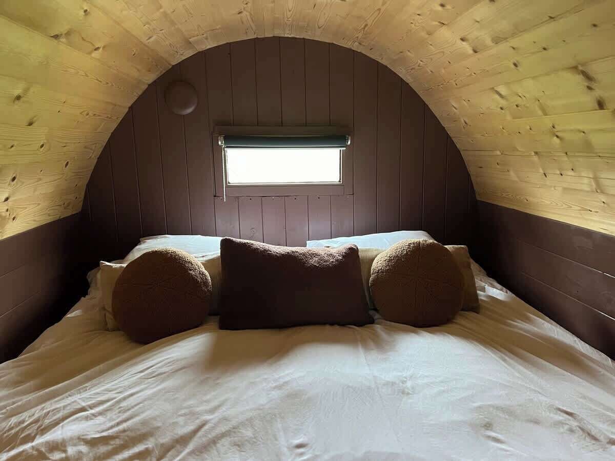 Une chambre mansardée chaleureuse avec un plafond en bois incurvé, une petite fenêtre et un lit bien fait, agrémenté d’oreillers.