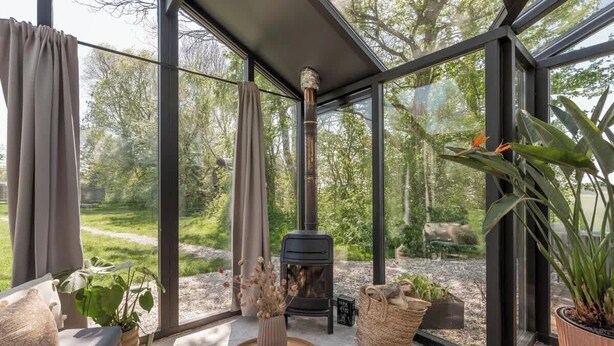 Ein moderner, abgeschlossener Raum mit Glaswand, Kaminofen und Blick auf eine bewaldete Landschaft.