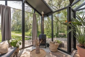 Ein moderner, abgeschlossener Raum mit Glaswand, Kaminofen und Blick auf eine bewaldete Landschaft.