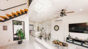 A modern living room with a white sofa, a dining table, and a TV. There is a staircase with orange steps leading to another level.