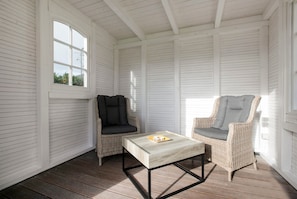 A room with white paneled walls, a wooden floor, a white table with a metal frame, and two wicker chairs with grey cushions.