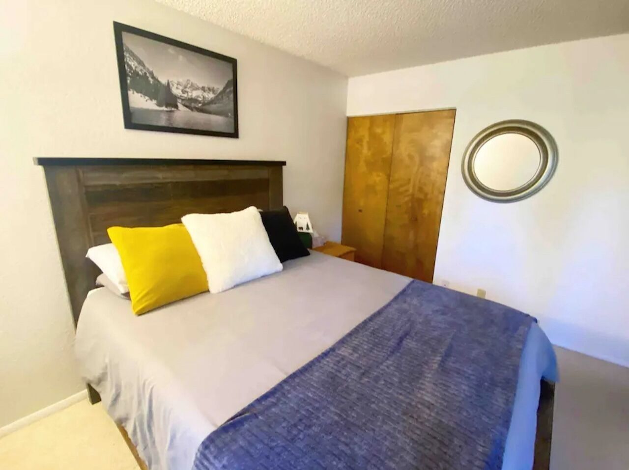 Una cama bien hecha con una manta gris y azul, un cabecero de madera y almohadas en tonos amarillo, blanco y negro.