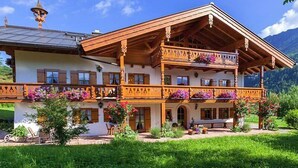 Un chalet traditionnel en bois, doté d’un balcon, entouré d’une végétation luxuriante et avec des montagnes en toile de fond.