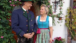 Un homme et une femme, vêtus de costumes traditionnels, se tiennent devant une porte surmontée d’une arche recouverte de fleurs.
