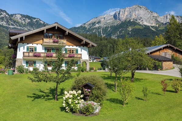 A two-story chalet with a balcony, surrounded by a well-maintained garden and a mountain backdrop.