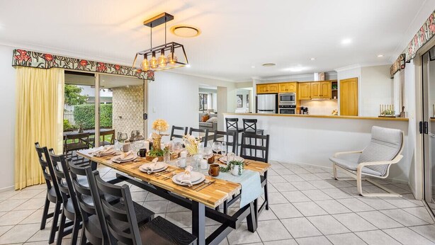 Un espace repas moderne avec une table en bois dressée pour six personnes, entourée de chaises noires. On aperçoit, au fond, une cuisine avec des meubles de couleur jaune.