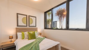 A bedroom with a bed, two bedside tables, a window with a view of palm trees and buildings, and two framed pictures on the wall. - One bed Apt at Southbank w/ Rooftop Pool&Carpark (South Brisbane)