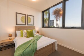 A bedroom with a bed, two bedside lamps, a window with a view of palm trees and buildings, and two framed pictures on the wall.