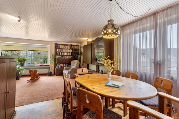 A cozy room with a wooden dining table, chairs, a bookshelf, and a large window with curtains.