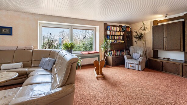 A living room with a beige sofa, a bookshelf, a chair, a wooden coffee table, and a window with a view of trees. - Holiday Home 'Haus Waldblick' with Private Terrace, Private Garden and Wi-Fi (Bad Berleburg)