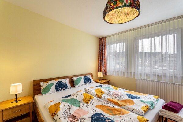 A hotel room with a bed, a nightstand, a lamp, a window with curtains, and a hanging light fixture.