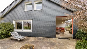 A house with a grey shingled exterior, a covered patio with a table and chairs, and a sun lounger. - Holiday Home 'Haus Waldblick' with Private Terrace, Private Garden and Wi-Fi (Bad Berleburg)
