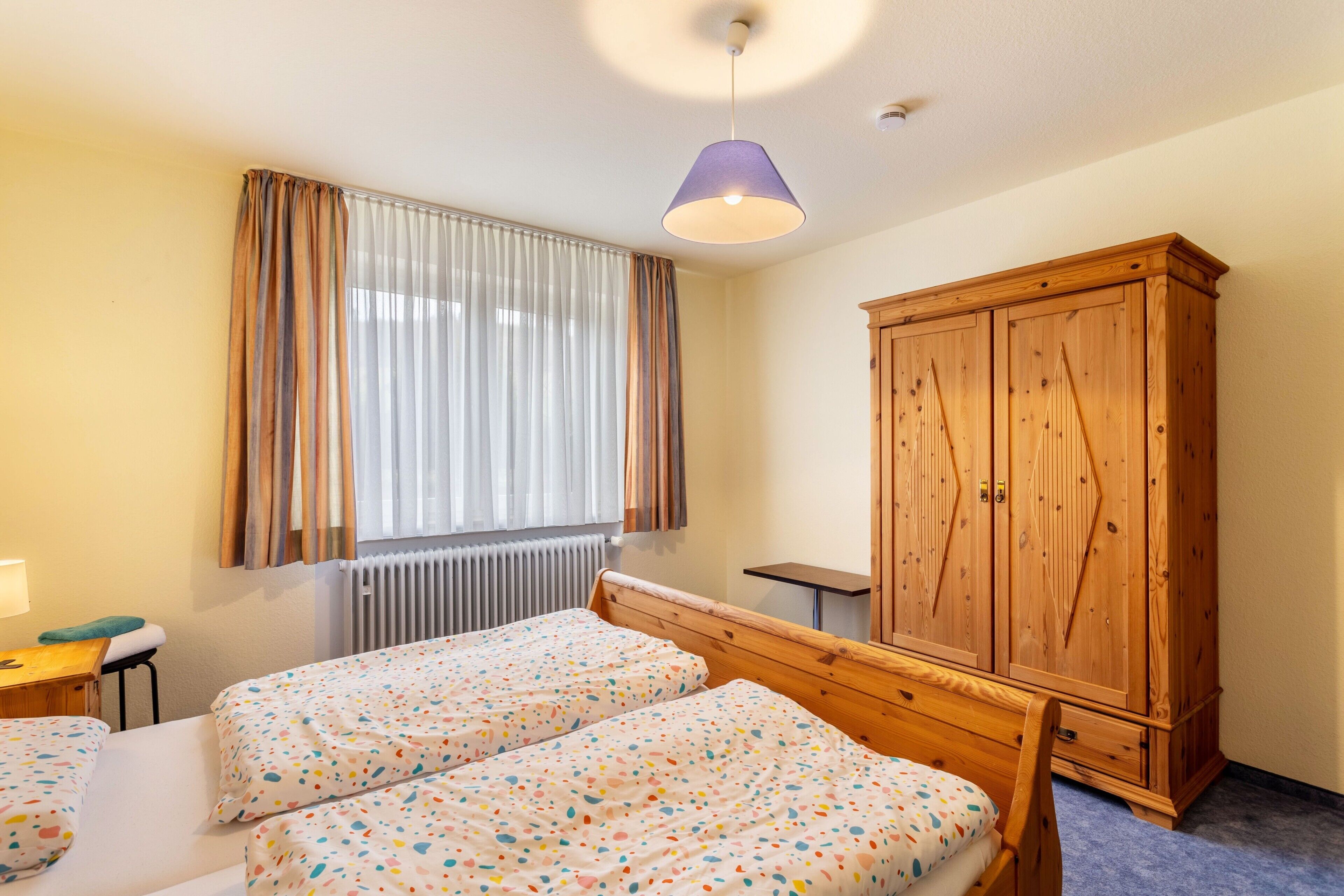 Une chambre avec deux lits, une armoire en bois, une fenêtre avec des rideaux et un luminaire au plafond.